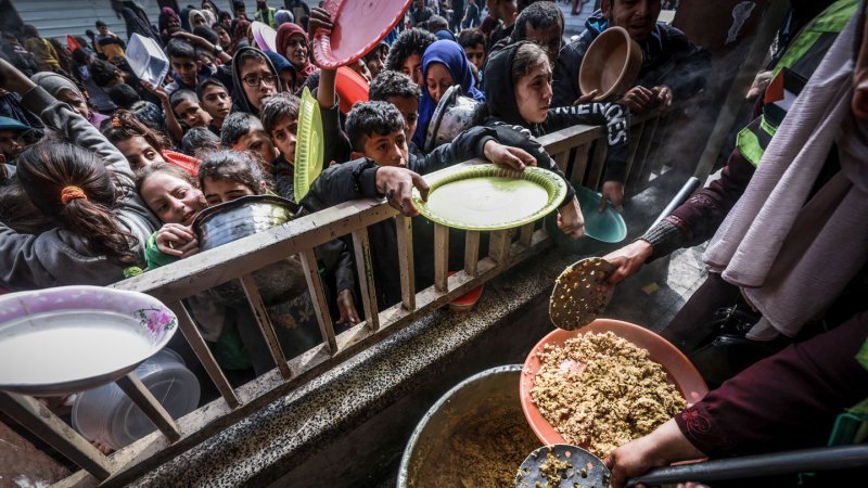 A large crowd of children stretch out plates and pots over a wooden railing toward a person dishing out food.