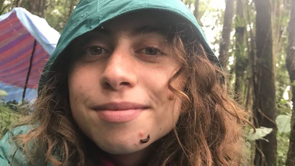 A closeup of researcher Mai Fahmy's face during an expedition to Madagascar to, in part, find leeches. She succeeded, as the one feeding on her chin attests. She's wearing a green hooded raincoat and a tent is visible behind her.