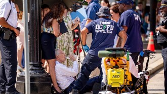 a group of people including EMTs huddle around an older white man drinking from a bottle of water while sitting on the ground in Washington D.C.